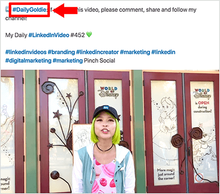 Этот снимок экрана показывает, как Голди Чан использует хэштеги в тексте своих видеопостов в LinkedIn. Красные выноски указывают на хэштег #DailyGoldie в тексте, который является уникальным для ее видеопостов и помогает ей отслеживать публикации. Сообщение также включает другие соответствующие хэштеги, которые помогают людям найти ее видео, в том числе #LinkedInVideo. На видеоизображении Голди стоит перед некоторыми дверями экспозиции World of Disney. Она азиатка с зелеными волосами. На ней черная кепка LinkedIn, черное колье-чокер, розовая рубашка с макаронным принтом и сине-белый пиджак.